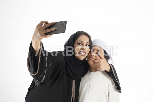 بورتريه لأم وإبن عربي خليجي إماراتي يرتديان الزي التقليدي ,  مستمتعتان بالتقاط صورة سيلفي من خلال الهاتف المحمول، التقاط صور تذكارية ، صورة جماعية ، توثيق اللحظات السعيدة ، خلفية بيضاء 