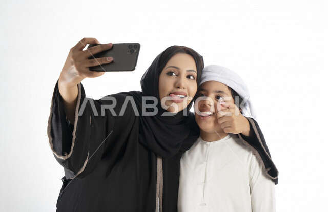 بورتريه لأم وإبن عربي خليجي إماراتي يرتديان الزي التقليدي ,  مستمتعتان بالتقاط صورة سيلفي من خلال الهاتف المحمول، التقاط صور تذكارية ، صورة جماعية ، توثيق اللحظات السعيدة ، خلفية بيضاء 