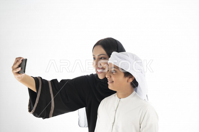 بورتريه لأم وإبن عربي خليجي إماراتي يرتديان الزي التقليدي ,  مستمتعتان بالتقاط صورة سيلفي من خلال الهاتف المحمول، التقاط صور تذكارية ، صورة جماعية ، توثيق اللحظات السعيدة ، خلفية بيضاء 