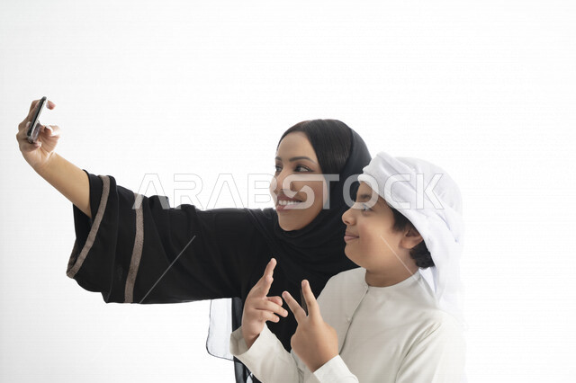 Portrait of an Emirati Arab mother and son in traditional dress, enjoying taking a selfie with their mobile phone, taking souvenir photos, a group photo, documenting happy moments, white background