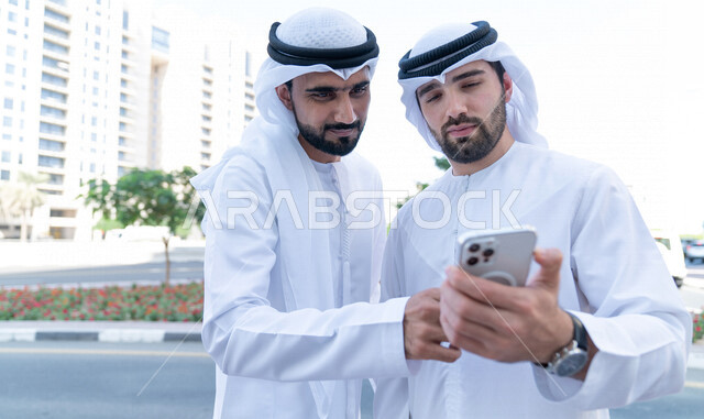 رجلان اعمال عربيان خليجييان اماراتييان يتصفحان الهاتف المحمول خارج مبنى الشركة، فريق عمل، مهن ووظائف اماراتية، محادثات ونقاشات عمل، زملاء عمل، إنتهاء الدوام، خروج الموظفين، التناقش بامور العمل، مناقشة وتنظيم الاعمال، تحقيق اهداف العمل