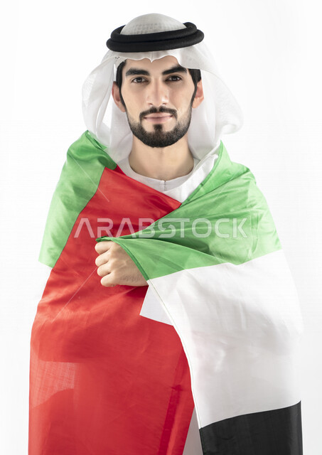 بورتريه لرجل عربي خليجي إماراتي، ينظر إلى الكاميرا، بالزي الإماراتي التقليدي، يحمل علم الإمارات العربية المتحدة على ظهره، الإحتفال باليوم الوطني الإماراتي، هوية اليوم الوطني الإماراتي 51 ، يوم استقلال الإمارات، حب الوطن، العيد الوطني الإماراتي