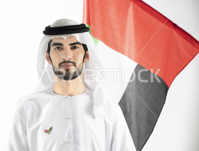 Portrait of an Emirati Arab Gulf man, looking at the camera, in traditional Emirati dress, against the background of the UAE flag, hanging the National Day emblem on his chest, celebrating the UAE National Day, the identity of the UAE National Day 51, UAE