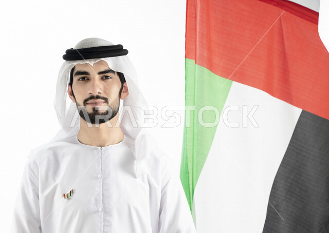 Portrait of an Emirati Arab Gulf man, looking at the camera, in traditional Emirati dress, against the background of the UAE flag, hanging the National Day emblem on his chest, celebrating the UAE National Day, the identity of the UAE National Day 51, UAE
