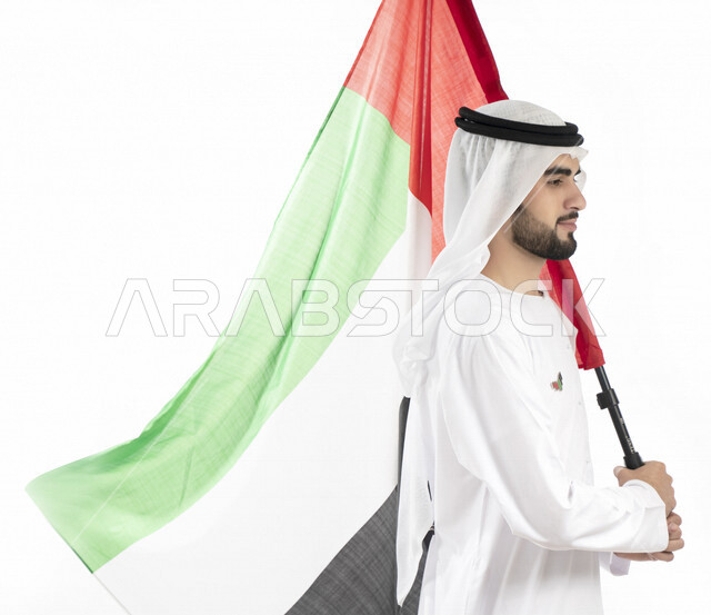 بورتريه زاوية من الجنب لرجل عربي خليجي إماراتي، يرتدي الزي الإماراتي التقليدي، يحمل علم الإمارات العربية المتحدة، تعليق شعار اليوم الوطني على صدره ، الإحتفال باليوم الوطني الإماراتي، هوية اليوم الوطني الإماراتي 51 ، يوم استقلال الإمارات، حب الوطن