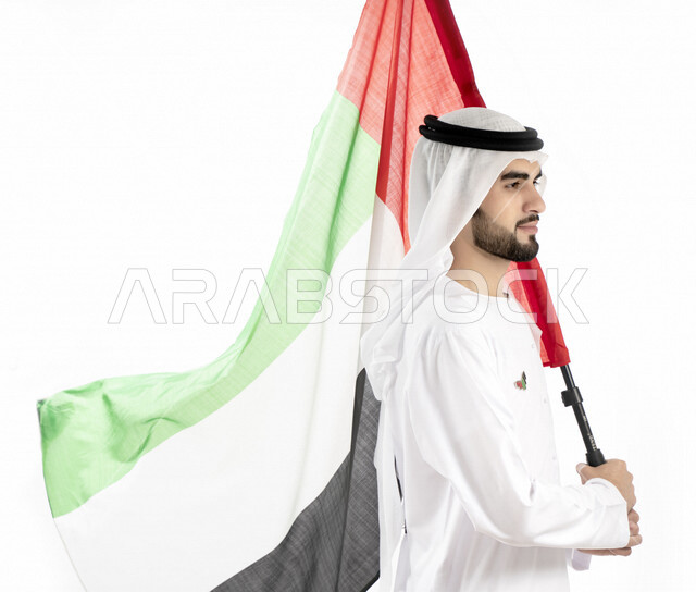 بورتريه زاوية من الجنب لرجل عربي خليجي إماراتي، يرتدي الزي الإماراتي التقليدي، يحمل علم الإمارات العربية المتحدة، تعليق شعار اليوم الوطني على صدره ، الإحتفال باليوم الوطني الإماراتي، هوية اليوم الوطني الإماراتي 51 ، يوم استقلال الإمارات، حب الوطن