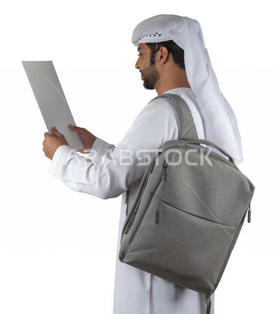 Portrait of an Arab Gulf Emirati university student with a bag on his back, holding a university book in his hand, reading university books, reviewing daily lessons, university education in the United Arab Emirates, white background.