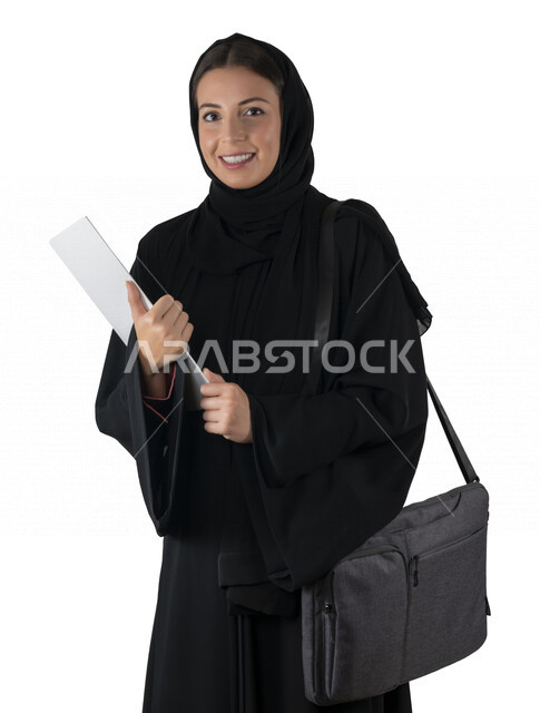 بورتريه لطالبة جامعية عربية إماراتيه خليجية محجبة مبتسمه تحمل حقيبة اللابتوب على كتفها, تمسك بيدها أوراق عمل, تنظر الى الكاميرا, موظفه في شركه إماراتيه, شابة متشوقة للذهاب الى العمل, خلفية بيضاء