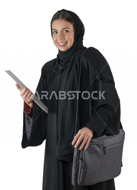 بورتريه لطالبة جامعية عربية إماراتيه خليجية محجبة مبتسمه تحمل حقيبة اللابتوب على كتفها, تمسك بيدها أوراق عمل, تنظر الى الكاميرا, موظفه في شركه إماراتيه, شابة متشوقة للذهاب الى العمل, خلفية بيضاء