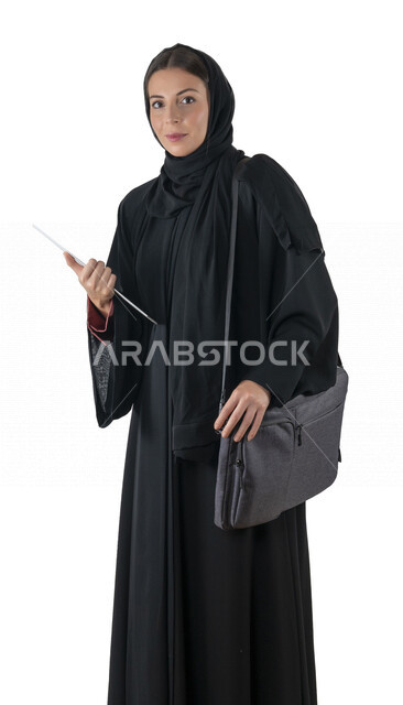 بورتريه لطالبة جامعية عربية إماراتيه خليجية محجبة مبتسمه تحمل حقيبة اللابتوب على كتفها, تمسك بيدها أوراق عمل, تنظر الى الكاميرا, موظفه في شركه إماراتيه, شابة متشوقة للذهاب الى العمل, خلفية بيضاء