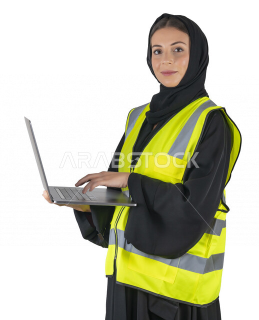 بورتريه لمهندسة معمارية عربية إماراتيه خليجية تحمل بيدها جهاز اللابتوب المحمول، تنظر الى الكاميرا, مهندسة إنشاءات محجبة ترتدي بدلة السلامة في العمل، مهن ووظائف هندسيه، مشروع بناء، مفهوم الهندسه المعماريه, خلفية بيضاء