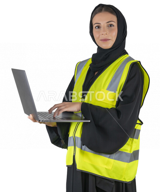 بورتريه لمهندسة معمارية عربية إماراتيه خليجية تحمل بيدها جهاز اللابتوب المحمول، تنظر الى الكاميرا, مهندسة إنشاءات محجبة ترتدي بدلة السلامة في العمل، مهن ووظائف هندسيه، مشروع بناء، مفهوم الهندسه المعماريه, خلفية بيضاء