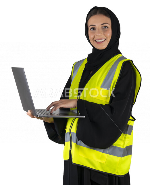بورتريه لمهندسة معمارية عربية إماراتيه خليجية تحمل بيدها جهاز اللابتوب المحمول، تنظر الى الكاميرا, مهندسة إنشاءات محجبة ترتدي بدلة السلامة في العمل، مهن ووظائف هندسيه، مشروع بناء، مفهوم الهندسه المعماريه, خلفية بيضاء