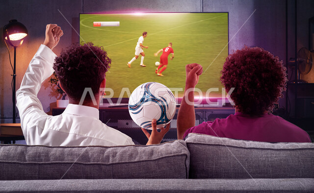 صورة من الخلف لشابان عربيان خليجيان سعوديان يستمتعان بمشاهدة مباريات كأس العالم على التلفاز، شاب يمسك بيده كرة القدم، ايماءات تدل على التشجيع والحماس، النجاح والقوة، تحقيق الاهداف، السعادة بالفوز،  كأس العالم 2022، مونديال قطر