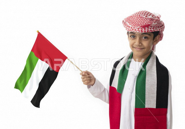 خلفية بيضاء, الإحتفال بيوم الاستقلال الوطني لدولة الإمارات العربية المتحدة, بورتريه مقرب لولد عربي خليجي اماراتي يرتدي الزي التقليدي الاماراتي يحمل بيده ويرتدي علم الإمارات, هوية اليوم الوطني الإماراتي51, تقديم الولاء والحب للوطن, يبتسم إلى الكاميرا