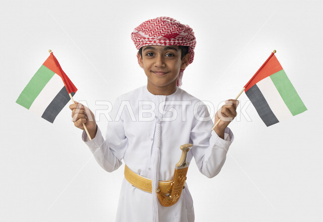 هوية اليوم الوطني الإماراتي51, الإحتفال بيوم الاستقلال الوطني لدولة الإمارات العربية المتحدة, بورتريه مقرب لولد عربي خليجي اماراتي يرتدي الزي التقليدي الاماراتي يحمل بيديه أعلام الإمارات, إيماءات الفرح والسعادة, تقديم الولاء والحب للوطن, خلفية بيضاء