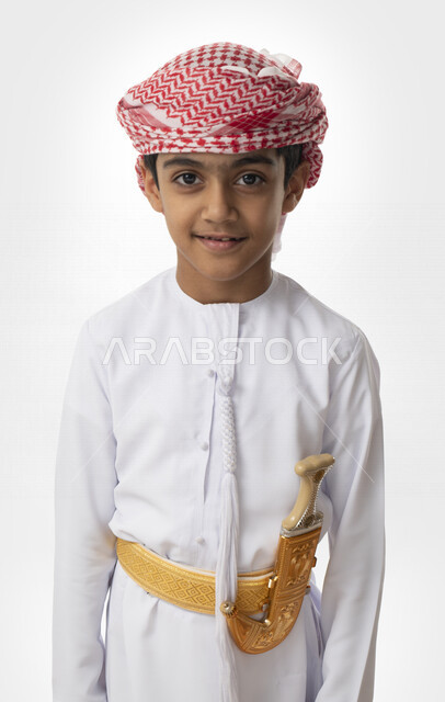الوقوف أمام الكاميرا مبتسم, ملابس وأزياء تراثية شعبية إماراتية تقليدية, بورتريه مقرب مقرب لولد عربي خليجي إماراتي يرتدي الكندوره والعمامه الاماراتية, إرتداء الحزام والخنجر الإماراتي الذهبي, خلفية بيضاء