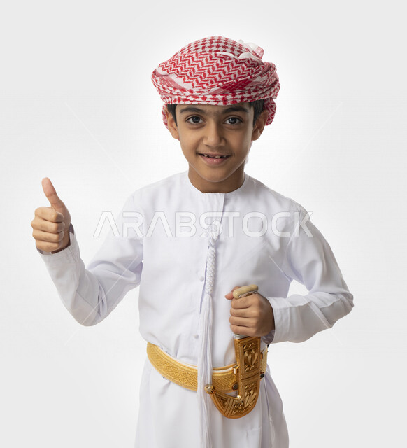 الإعجاب والرضا بالنتيجة, ملابس وأزياء تراثية شعبية إماراتية تقليدية, بورتريه مقرب لولد عربي خليجي إماراتي يرتدي الكندوره والعمامه الاماراتية, يمسك بيده الخنجر الاماراتي الذهبي, إيماءات وجه ويدين تدل على الموافقة والقبول, خلفية بيضاء