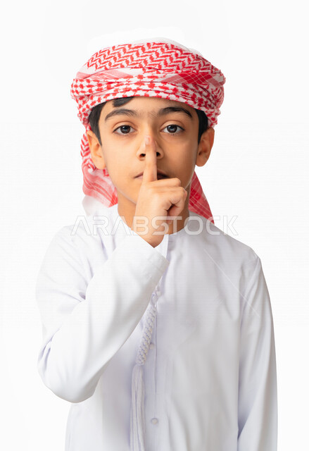 التوقف عن الكلام و الأمر بالصمت والسكوت، بورتريه لطفل عربي سعودي اماراتي يرتدي االكندورة والعمامة الاماراتية ويرفع اصبعه الى فمه, إيماءات يدين تدل على طلب الهدوء و عدم الإزعاج, خلفية بيضاء