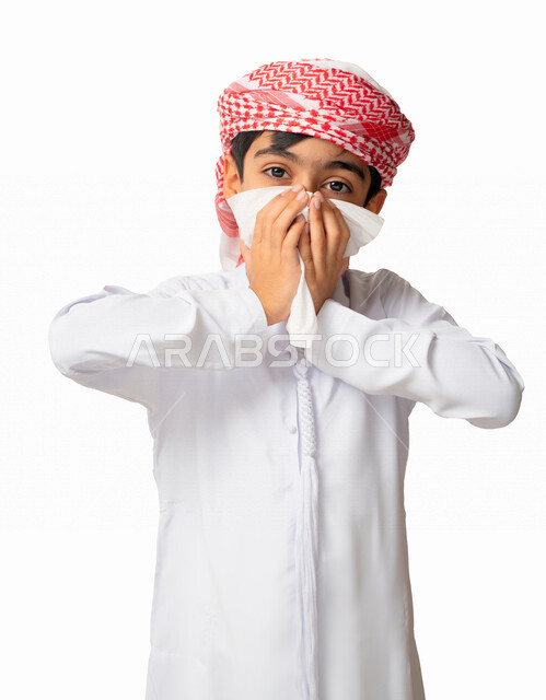 Looking at the camera, the need for medicine and treatment, feeling sick and having a cold, allergic rhinitis, using tissues for sneezing, a portrait of an Arab Emirati Gulf boy wearing a turban and a kandora with face and hands gestures indicating sneezi