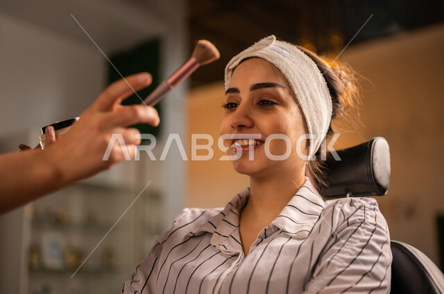 مرأة عربية خليجية سعودية تضع إحدى أدوات التجميل (الفرشاة) على بشرة الوجه بيد فنانة مكياج في صالون تجميلي نسائي، مرأة خبيرة بمستحضرات التجميل، مفهوم الإهتمام بالجمال والمظهر الخارجي، ادوات مكياج تتوريال، التسويق لمنتجات التجميل