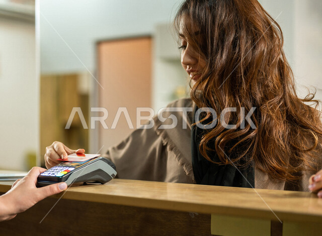 امرأة عربية خليجية سعودية تقوم بالدفع عند الإستقبال بطريقة الكترونية بإستخدام بطاقة الإئتمان البنكية في داخل الصالون التجميلي النسائي ، تعابير تدل على السعادة ، مفهوم الإهتمام بالجمال والمظهر الخارجي