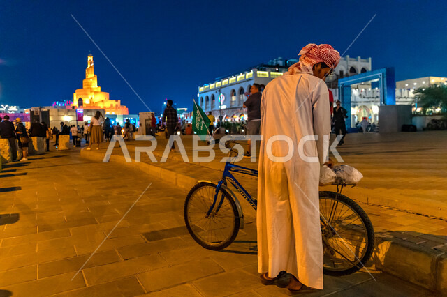 صورة من الخلف لرجل عربي خليجي مع دراجته الهوائية، مجموعة من الجماهير خارج احد استادات قطر، كأس العالم في قطر 2022، بطولة كأس العالم، فيفا 2022، مونديال قطر، السياحة في قطر