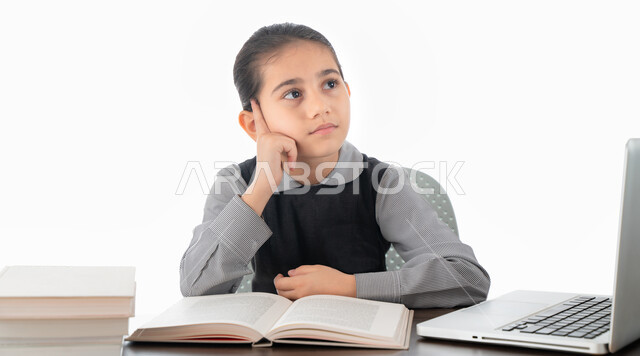 اللهو أثناء الدراسة، ايماءات وجه ويدين تدل على التفكير بشيء ما، سلبيات التعليم عن بعد، بورتريه لتلميذة عربية خليجية اماراتية ترتدي الزي المدرسي تجلس على طاولة الدراسة أمام جهاز الكمبيوتر، طالبة سعودية تنظر إلى شيء ما، خلفية بيضاء