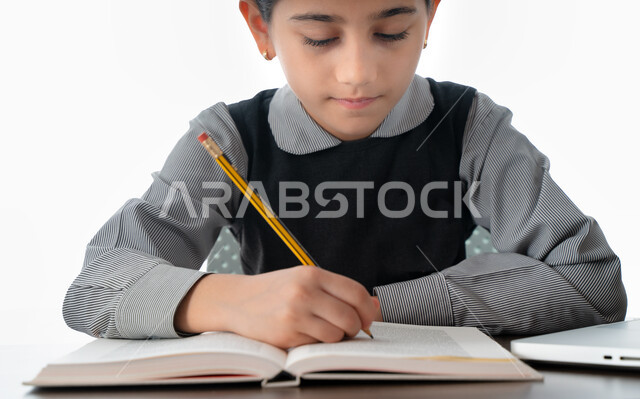 التحضير الجيد للإختبارات، الجلوس على طاولة الدراسة لمراجعة الدروس، الاجتهاد والتفوق ومتابعة الدراسة، بورتريه لتلميذة عربية خليجية اماراتية ترتدي المريول المدرسي تقوم بكتابة الوظائف المنزلية، طالبة سعودية تدرس بتركيز وتمعن، خلفية بيضاء