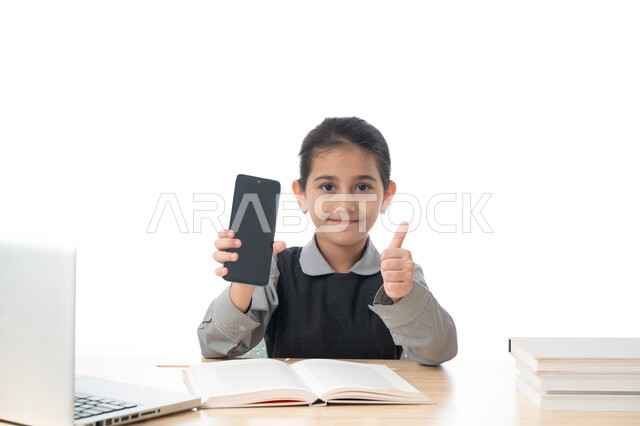 Distance learning using a computer, a portrait of an Arab Gulf Emirati student wearing a school uniform, holding a mobile phone in her hand, raising her thumb up, a smiling Saudi student looking at the camera, face and hands gestures indicating acceptance