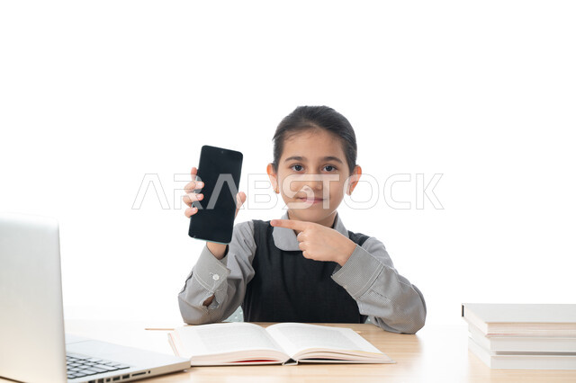 مفهوم التعلم عن بعد باستخدام وسائل التكنولوجيا الحديثة، بورتريه لطالبة عربية خليجية اماراتية ترتدي المريول المدرسي تمسك بيدها الجوال، تلميذة سعودية مبتسمة تشير إلى الهاتف المحمول، عرض شاشة سوداء، الدراسة من خلال منصات التعليم، خلفية بيضاء