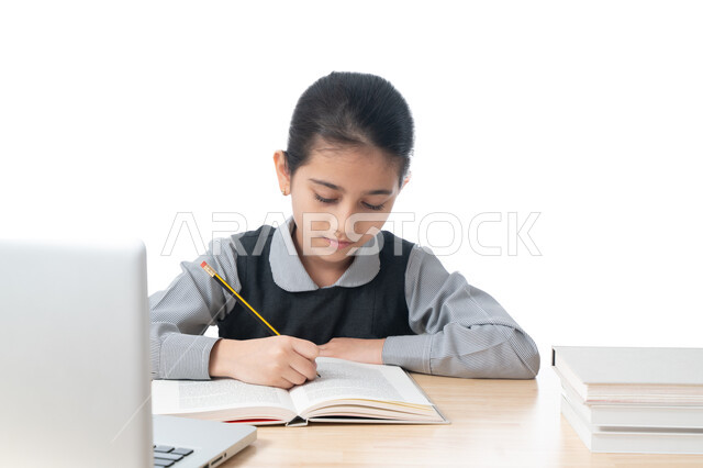 متابعة الدراسة عن طريق المنصات الدراسية الإلكترونية، بورتريه لطالبة عربية خليجية اماراتية ترتدي الزي المدرسي تقوم بتحضير الدروس باستخدام الكمبيوتر، تلميذة سعودية مجتهدة تجلس على طاولة الدراسة وتكتب واجباتها، مفهوم التعليم الإلكتروني، خلفية بيضاء