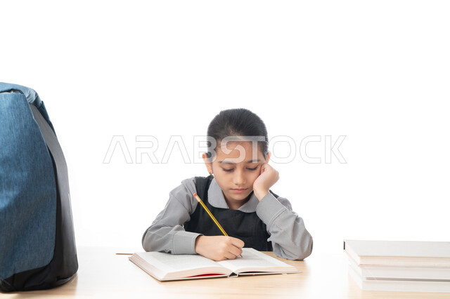 الجلوس على طاولة الدراسة لمراجعة الدروس، الاجتهاد والتفوق ومتابعة الدراسة، بورتريه لتلميذة عربية خليجية اماراتية ترتدي المريول المدرسي تقوم بكتابة الوظائف المنزلية، طالبة سعودية تدرس بتركيز وتمعن، التحضير الجيد للإختبارات، خلفية بيضاء