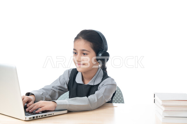 التعلم عن بعد باستخدام وسائل التكنولوجيا الحديثة، بورتريه لطالبة عربية خليجية اماراتية ترتدي المريول المدرسي وسماعات الرأس تقوم بمتابعة الدروس باستخدام الكمبيوتر، تلميذة سعودية مبتسمة تجلس على طاولة الدراسة، مفهوم التعليم الإلكتروني، خلفية بيضاء
