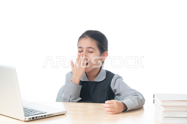 ايماءات وجه ويدين تدل على النعاس و التعب والإرهاق، بورتريه لطالبة عربية خليجية اماراتية ترتدي اليونيفورم المدرسي تقوم بالتثاؤب، سلبيات التعلم عن بعد، تلميذة سعودية تجلس على طاولة الدراسة وتشعر بالكسل والخمول، الحاجة إلى النوم والراحة، خلفية بيضاء