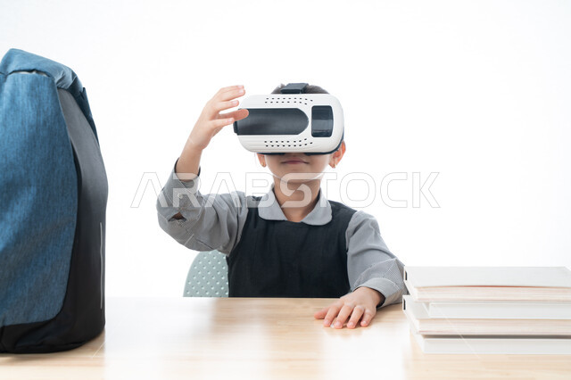 استخدام نظارات الواقع الافتراضي في التعليم، بورتريه لتلميذة عربية خليجية اماراتية ترتدي المريول المدرسي تشير إلى شيء ما وتتفاعل مع العالم الافتراضي التعليمي، طالبة سعودية تجلس على طاولة الدراسة، جمع التعلم مع الترفيه بواسطة التقنية الحديثة، خلفية بيضاء