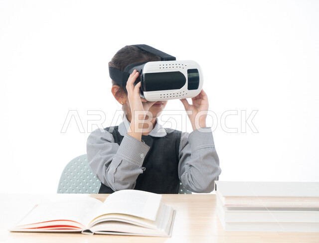 اكاديميات ومدارس حديثة و متطورة، جمع التعلم مع الترفيه باستخدام التقنية والتكنولوجيا الحديثة، بورتريه لتلميذة عربية خليجية اماراتية ترتدي الزي المدرسي تمسك بيديها نظارات العالم الافتراضي التعليمي، طالبة سعودية تجلس على طاولة الدراسة، خلفية بيضاء