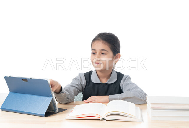 متابعة الدروس عن طريق منصات التعلم عن بعد، بورتريه لطالبة عربية خليجية اماراتية ترتدي الزي المدرسي تتابع دروسها باستخدام جهاز التابلت التقني، طرق تعليمية جديدة، تلميذة سعودية تجلس على طاولة الدراسة تقوم بالنقر على شاشة التابلت الحديث، خلفية بيضاء
