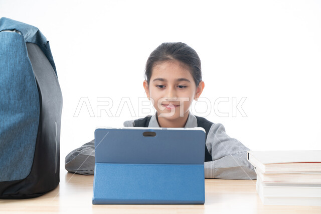 The concept of e-learning, distance education using modern technology, advanced learning methods, a portrait of an Arab Gulf Emirati student wearing a school apron following lessons using a technical tablet, a smiling Saudi schoolgirl sitting at the study
