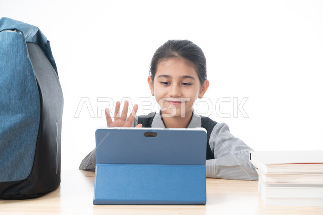 المشاركة الفعالة للطلاب ومتابعة الدروس والواجبات من خلال منصات التعلم عن بعد، بورتريه لتلميذة عربية خليجية اماراتية ترتدي الزي المدرسي تحضر دروسها عن طريق جهاز التابلت الإلكتروني، طالبة سعودية تجلس على طاولة الدارسة ترفع يدها للإجابة، خلفية بيضاء