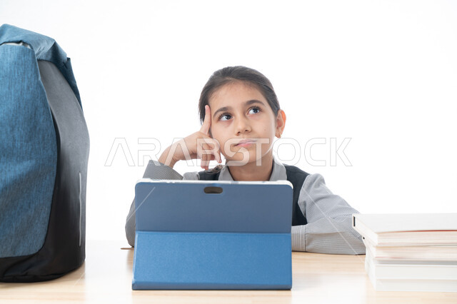 ايماءات وجه ويدين تدل على التفكير بشيء ما، استخدام التقنية الحديثة في التعليم، بورتريه لتلميذة عربية خليجية اماراتية ترتدي الزي المدرسي تتابع دروسها عن طريق جهاز التابلت الإلكتروني، طالبة سعودية تجلس على طاولة الدارسة وتنظر إلى شيء ما، خلفية بيضاء