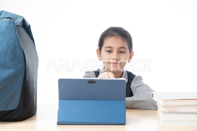 استخدام التقنية والتكنولوجيا الحديثة في التعليم، بورتريه لتلميذة عربية خليجية اماراتية ترتدي المريول المدرسي تقوم بمتابعة دروسها بواسطة جهاز التابلت الإلكتروني، مفهوم الدراسة عن بعد، طالبة سعودية تجلس على طاولة الدارسة وتشير لشيء ما، خلفية بيضاء