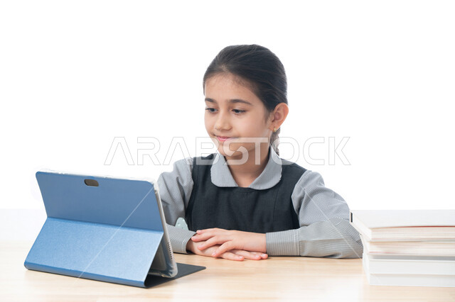 التعليم عن بعد باستخدام التكنولوجيا الحديثة، طرق تعلمية جديدة ومتطورة، بورتريه لطالبة عربية خليجية اماراتية ترتدي المريول المدرسي تقوم بمتابعة الدروس باستخدام جهاز التابلت التقني، تلميذة سعودية تجلس على طاولة الدراسة، مفهوم التعليم الإلكتروني، خلفية بيضاء