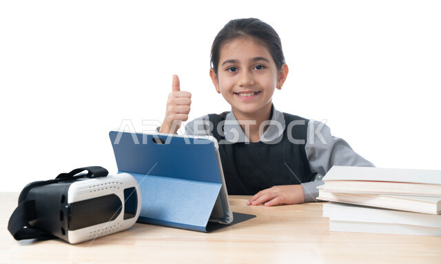 التعلم عن بعد باستخدام جهاز التابلت، بورتريه لطالبة عربية خليجية اماراتية ترتدي الزي المدرسي و بجانبها نظارات الواقع الافتراضي ترفع اصبع الابهام للأعلى، تلميذة سعودية مبتسمة تنظر إلى الكاميرا، ايماءات وجه ويدين تدل على والقبول والموافقة، خلفية بيضاء
