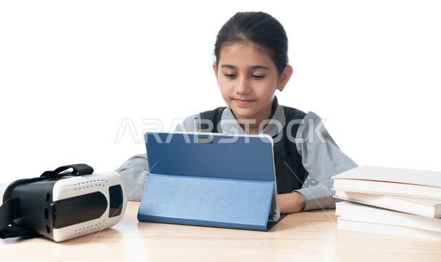 متابعة حصص الاونلاين عن طريق جهاز التابلت، بورتريه لطالبة عربية خليجية اماراتية ترتدي اليونيفووم المدرسي تضع بجانبها نظارات الواقع الافتراضي والكتب، التعلم عن بعد باستخدام التكنولوجيا الحديثة، تلميذة سعودية تجلس على طاولة الدراسة، خلفية بيضاء
