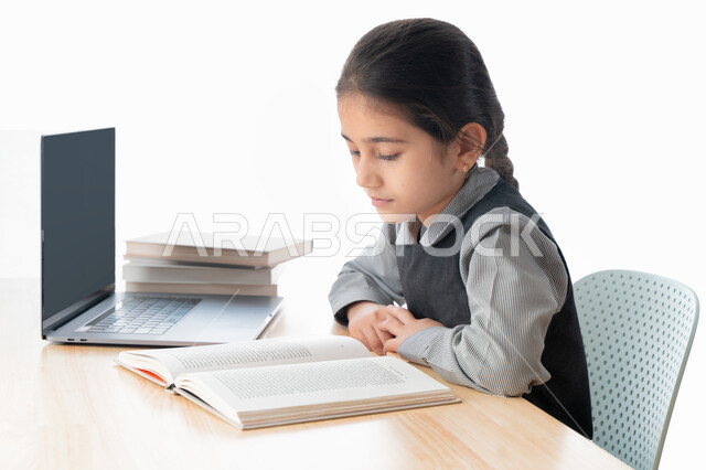 متابعة الحصص و الواجبات عبر الانترنت،  بورترية مقرب لتلميذة خليجية عربية إماراتية ترتدي مريول المدرسة و تدرس في المنزل، المذاكرة و الدراسة عن بعد، مفهوم التعليم الإلكتروني، طالبة سعودية مجتهدة تراجع دروسها من الكتب وتستخدم الحاسوب المحمول، خلفية بيضاء