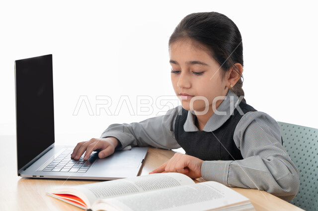 Studying and studying from a distance, a close-up portrait of a Gulf Arab Emirati student wearing a school apron and studying at home, the concept of e-learning, a diligent Saudi student reviewing her lessons from books and using a laptop, following up on