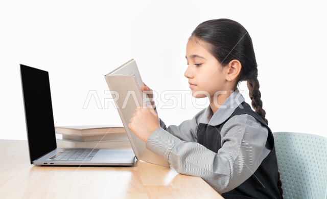 متابعة الحصص و حل الواجبات عبر الانترنت، بورترية مقرب لتلميذة خليجية عربية إماراتية ترتدي مريول المدرسة و تدرس في المنزل، المذاكرة و الدراسة عن بعد، مفهوم التعليم الإلكتروني، طالبة سعودية مجتهدة تراجع دروسها من الكتب وتستخدم الحاسوب المحمول، خلفية بيضاء