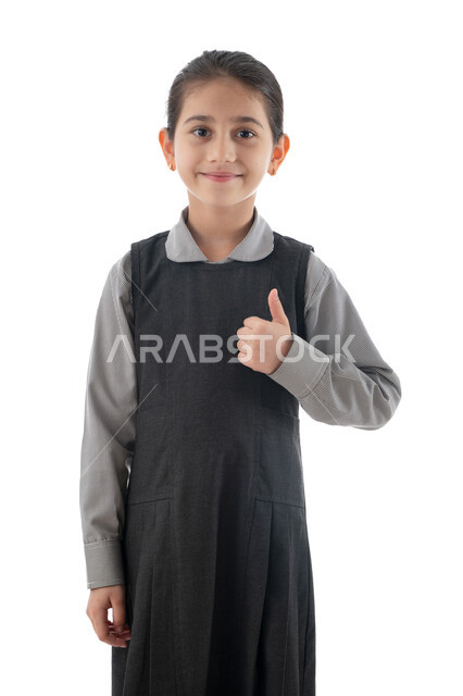 فصل تعليمي جديد وتحضيرات بداية العام الدراسي, بورتريه لطالبة عربية خليجية إماراتية مبتسمة ترتدي اليونيفوم المدرسي تنظر إلى الكاميرا, تلميذة سعودية تشير بإصبعها الإبهام للأعلى بإيماءات تدل على الحماس والمثابرة على الدراسة لتحقيق النجاح, خلفية بيضاء