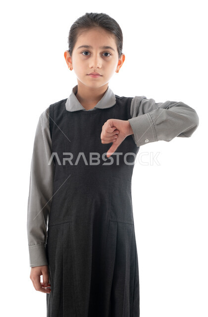 التنمر ومشاكل الأطفال مع أقرانهم في المدارس, ايماءات الرفض وعدم الرغبة بالذهاب إلى المدرسة, بورتريه لتلميذة عربية خليجية إماراتية ترتدي اليونيفورم المدرسي تنظر إلى الكاميرا, طالبة سعودية تشير بإصبعها الإبهام للأسفل وتشعر بالحزن لعودة الدراسة, خلفية بيضاء